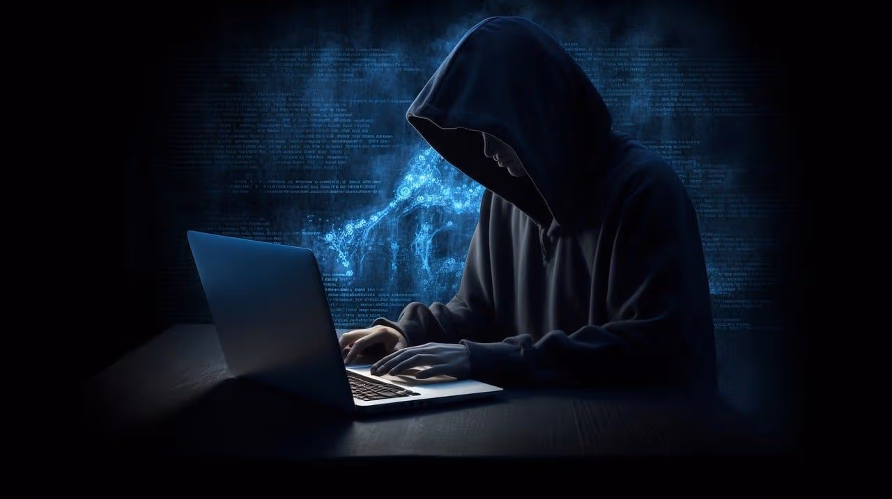 A hooded figure sits and types on a laptop in a dark room. (Pixabay)